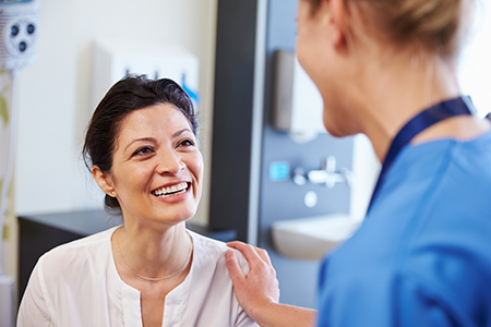 The image shows a woman sitting in a dental chair with her mouth open, smiling at the camera, while a dentist stands behind her, appearing to be engaged in conversation or examination.