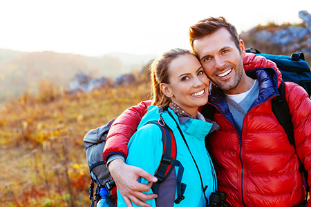 A man and woman are standing close together in an outdoor setting, smiling at the camera, with the man wearing a backpack.