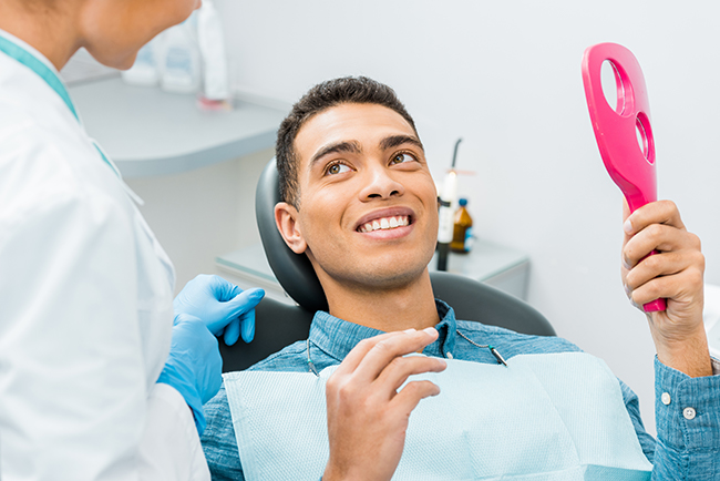 The image shows a smiling man seated in a dental chair, holding a pink object that appears to be an electric toothbrush, with a dental hygienist standing behind him wearing protective gloves and a face mask.