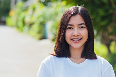 A young woman with short hair smiles at the camera, wearing a white top against a blurred background featuring greenery and a pathway.