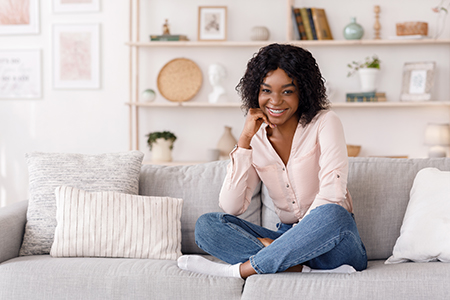 A woman sitting on a couch with a smile, wearing a pink blouse and blue jeans.